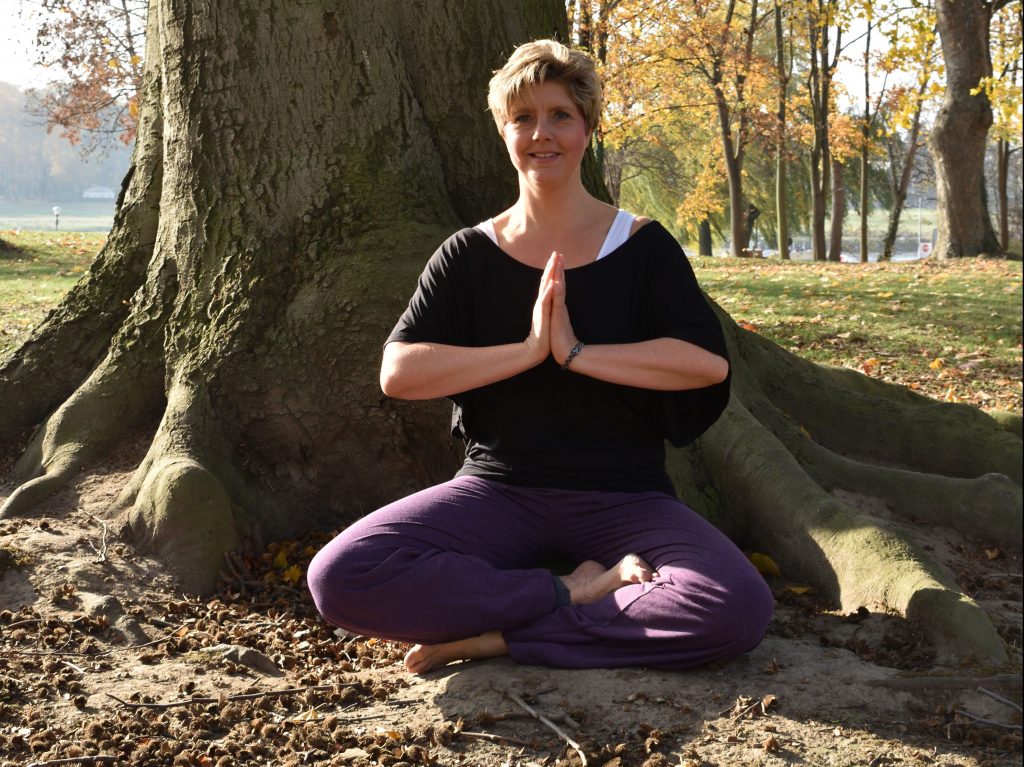 Mein Name ist Claudia Gründer und ich sitze im Yoga Schneidersitz vor einem großen Baum.